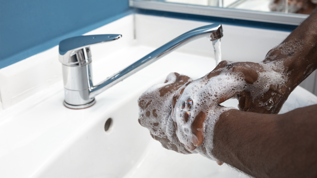 Hands lathering with CBD soap under a stream of water at a white sink with a chrome faucet. The soap creates a foamy lather, and the background features a blue wall and a mirror in a modern bathroom setting