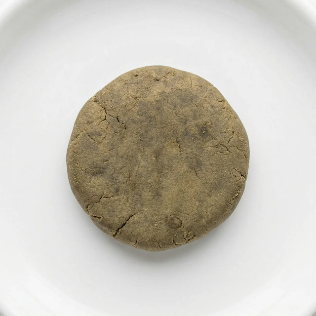 THCA bubble hash disc — warm olive-brown cured exterior with natural stress fractures, overhead view on white ceramic