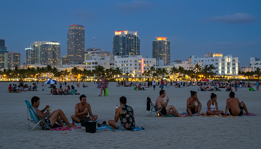 A diverse group on a Miami Beach cannabis-friendly section at dusk, using Delta-8 vapes and CBD drinks, with the skyline and art-deco buildings in the background.