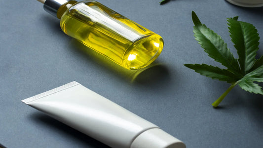 A CBD skincare and wellness product arrangement featuring a glass dropper bottle with golden CBD oil, a white cosmetic tube, a cream jar, and cannabis leaves on a dark surface | Hurcann