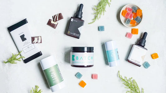 A flat-lay display of CBD wellness products, including oils, balms, gummies, and chocolate bars, arranged on a light background with green sprigs for a fresh, natural aesthetic | Hurcann