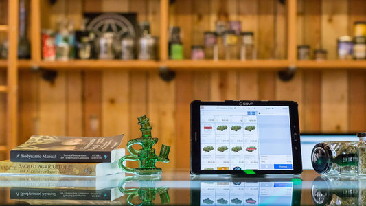 A cannabis dispensary counter featuring a glass surface with books, a green glass bong, a tablet displaying a cannabis product catalog, and jars of cannabis | Hurcann