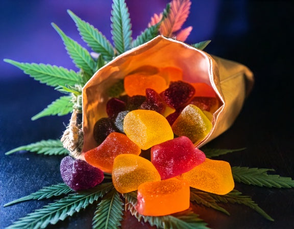 Terpene-enriched gummies in orange, purple, and green spilling from a hemp pouch with terpene wisps and a cannabis flower backdrop.