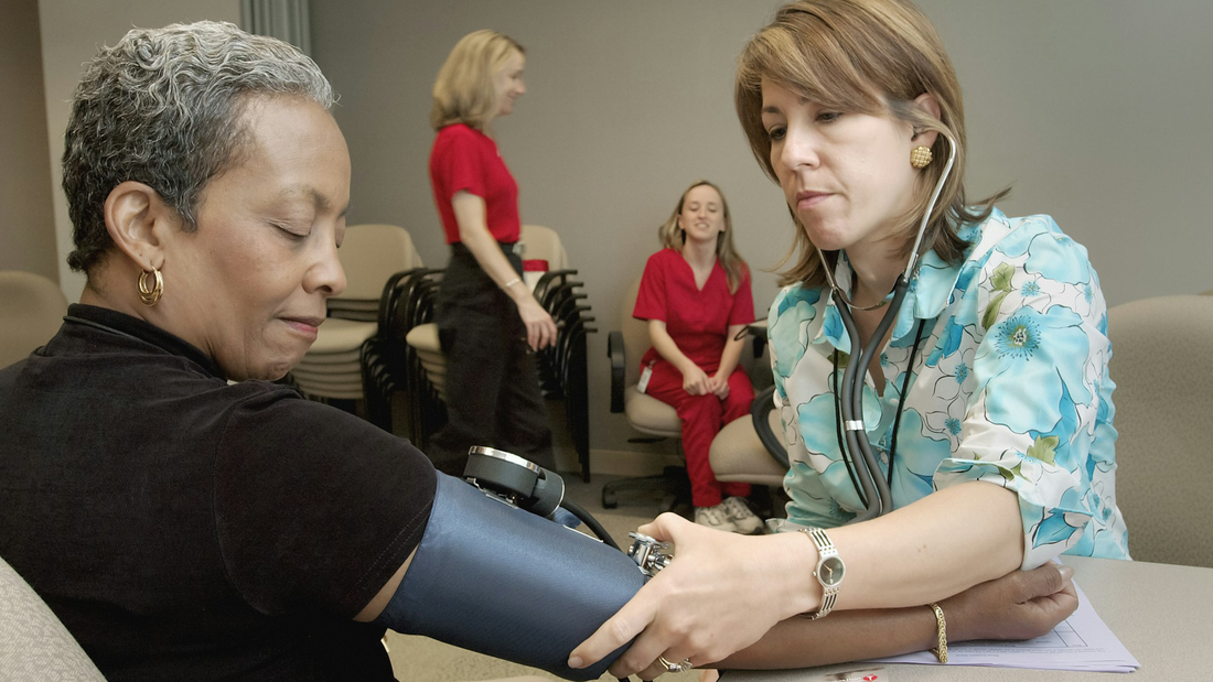 A healthcare professional in a blue floral blouse uses a sphygmomanometer to check an older woman's blood pressure in a medical facility. In the background, two other medical staff members in red uniforms are engaged in conversation