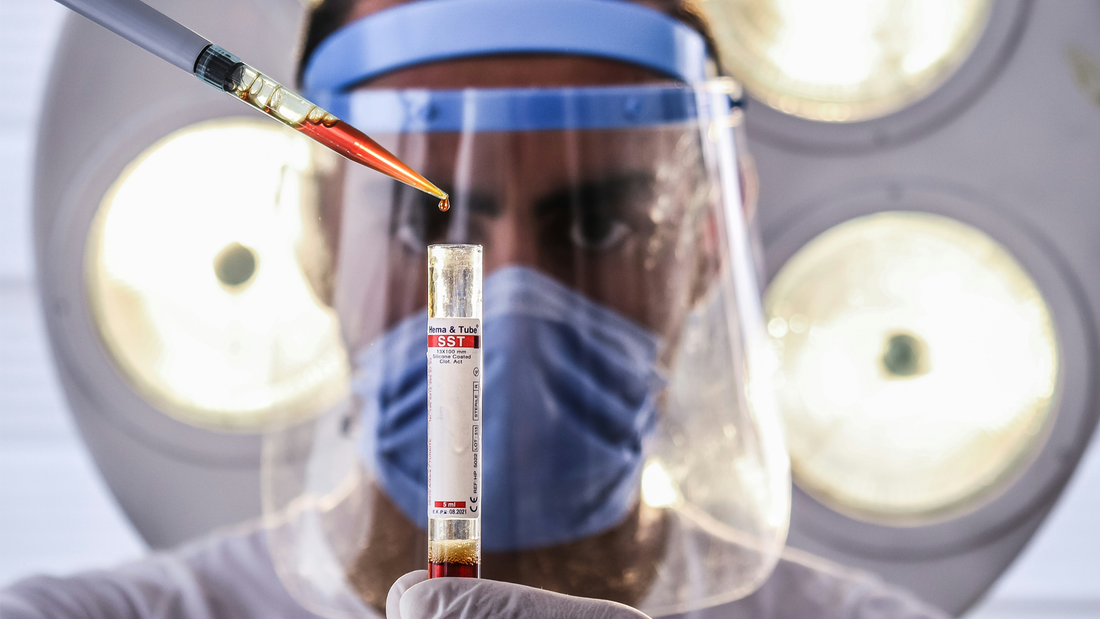 A scientist wearing a face shield, mask, and gloves carefully examines a test tube filled with a blood sample while using a pipette to add a liquid substance, with bright surgical lights in the background