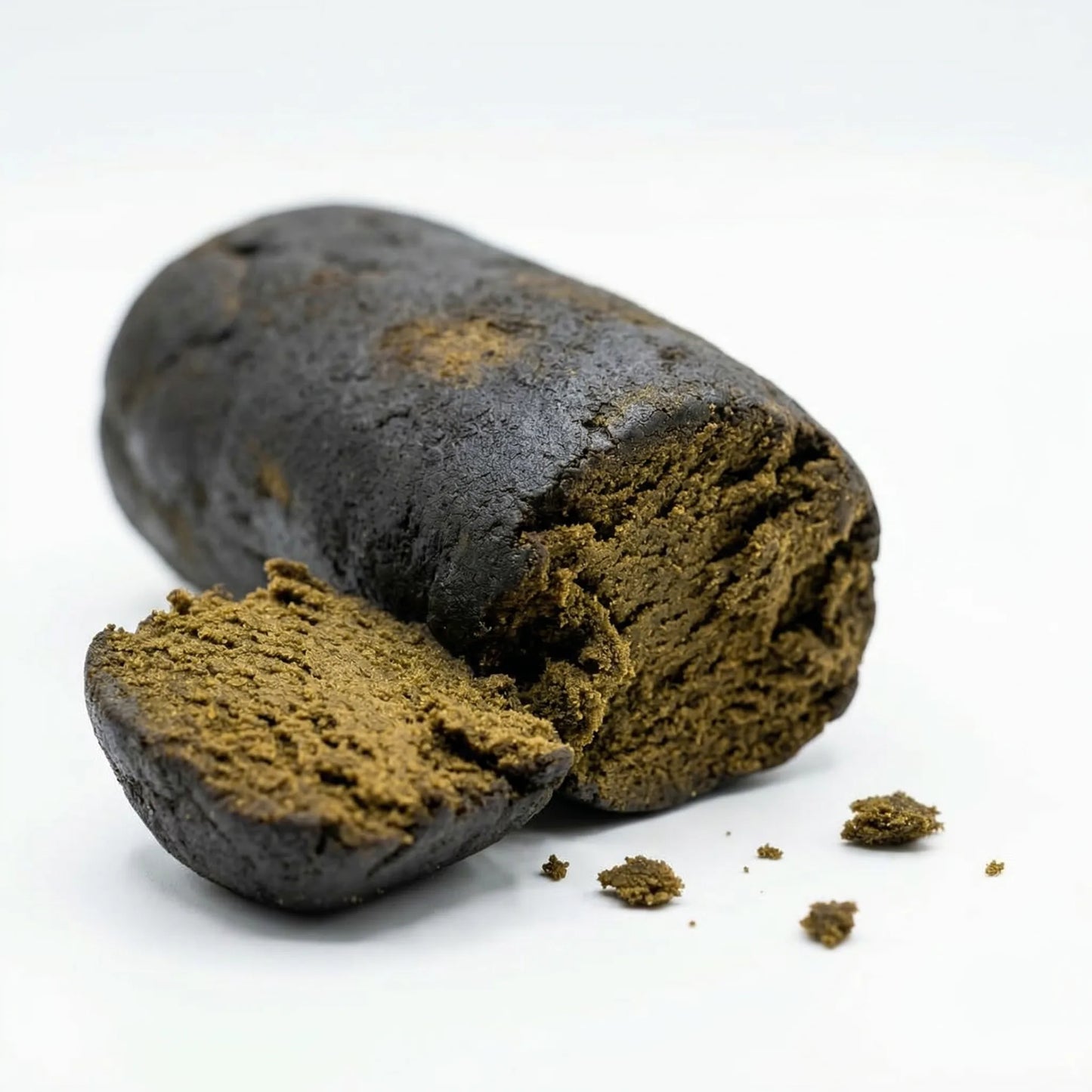 Bulk wholesale black hash log broken open revealing warm olive-brown granular interior, near-black exterior with slightly glossy surface, fine crumble scattered on white background — wholesale hemp hash concentrate bulk supply