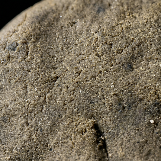 Extreme macro close-up of CBD bubble hash surface revealing dense sandy trichome texture with subtle sparkle in warm olive-brown tones
