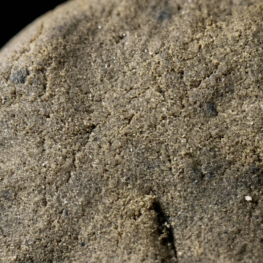 Extreme macro close-up of CBD bubble hash surface revealing dense sandy trichome texture with subtle sparkle in warm olive-brown tones | Hurcann