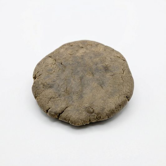 Round pressed disc of CBD bubble hash on a white background, showing an olive-brown surface with a fine cracked texture