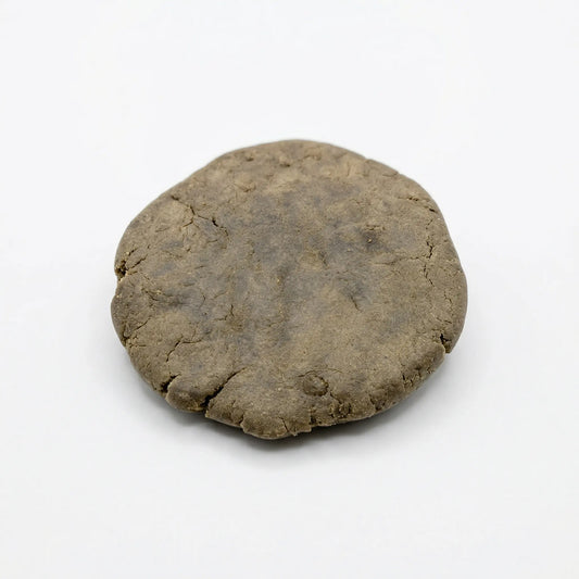 Round pressed disc of CBD bubble hash on a white background, showing an olive-brown surface with a fine cracked texture | Hurcann