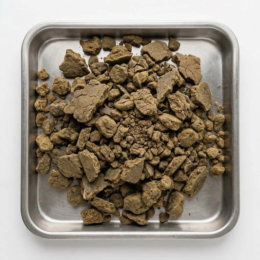 Overhead view of bulk CBD bubble hash chunks and crumbled pieces filling a square stainless steel tray, olive-brown tones with matte cracked texture, clean white background | Hurcann