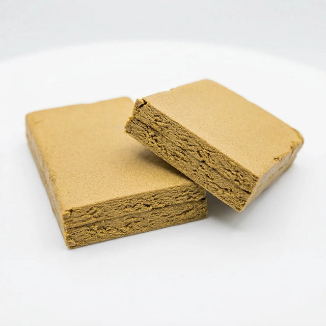 Two THCA La Mousse hash slabs on a white background, warm golden-tan smooth matte surface, one slab lying flat with a second slab leaning against it at an angle revealing a thick layered fibrous interior with compressed horizontal striations running through the full depth of the slab