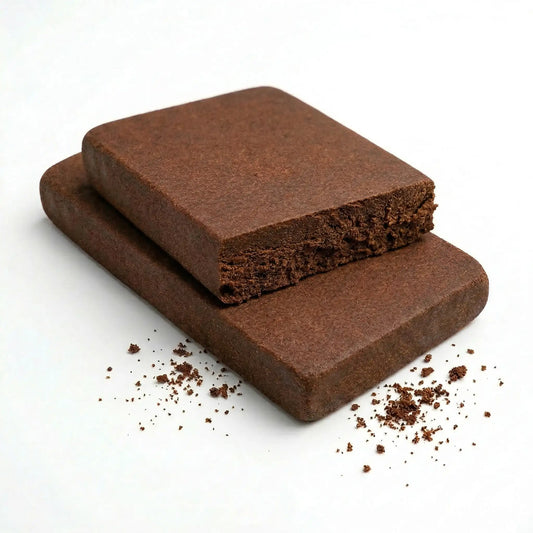 Two bulk wholesale THCA Lebanese red hash slabs stacked on a white background, top slab broken open revealing dense dark granular interior, smooth chocolate-brown matte exterior on both slabs, fine crumble scattered around the base — wholesale THCA Lebanese hash bulk concentrate