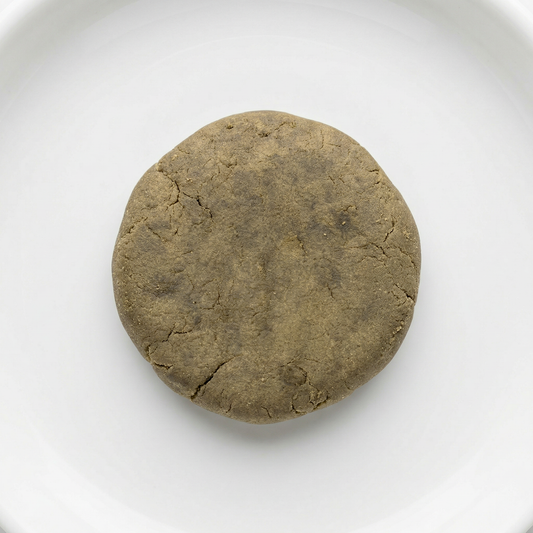 THCA bubble hash disc — warm olive-brown cured exterior with natural stress fractures, overhead view on white ceramic