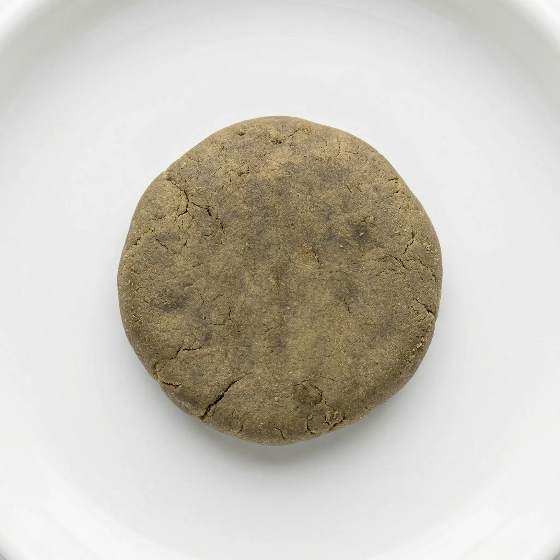 THCA bubble hash disc — warm olive-brown cured exterior with natural stress fractures, overhead view on white ceramic