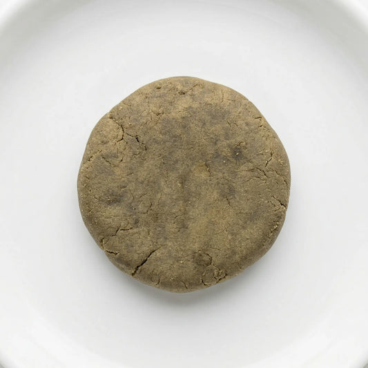 THCA bubble hash disc — warm olive-brown cured exterior with natural stress fractures, overhead view on white ceramic