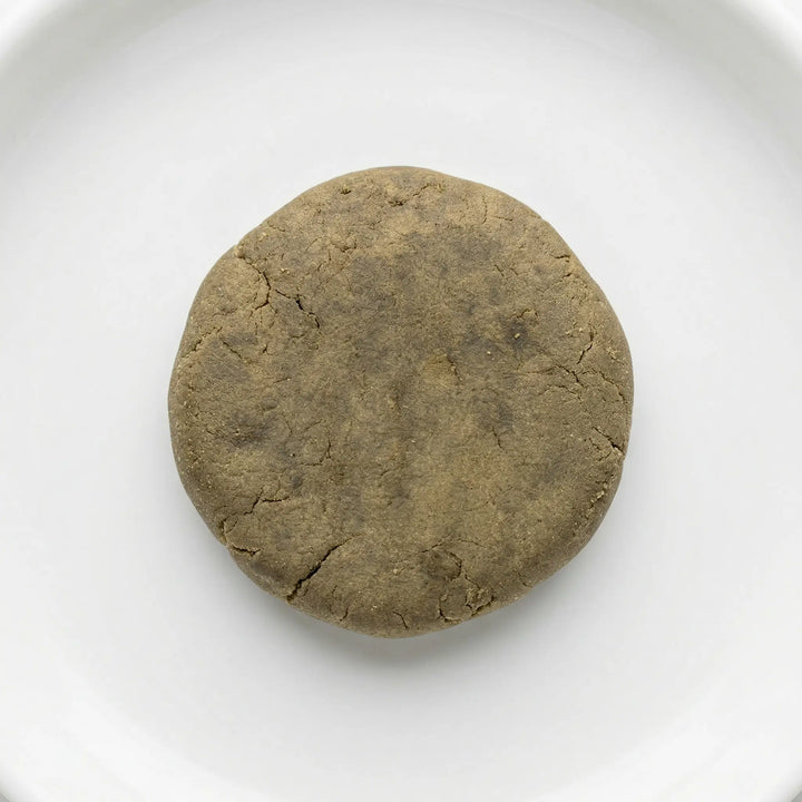 THCA bubble hash disc — warm olive-brown cured exterior with natural stress fractures, overhead view on white ceramic