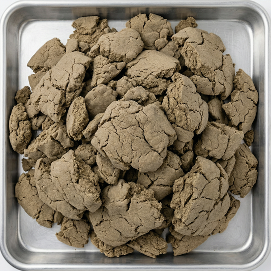 Overhead view of bulk THCA bubble hash chunks piled in a stainless steel tray, showing olive-brown cracked texture across multiple broken pieces