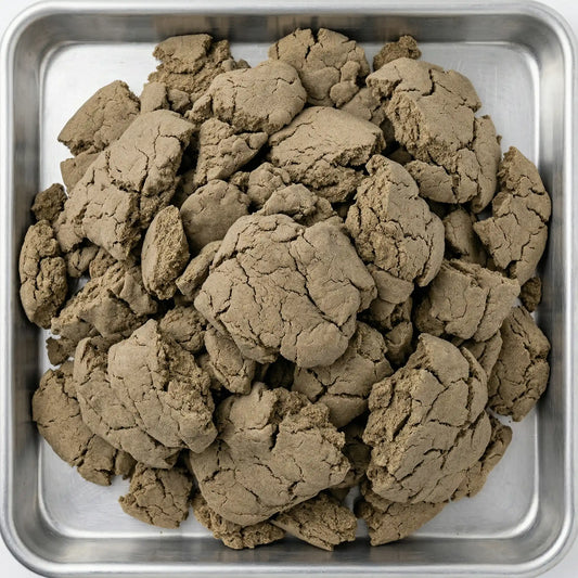 Overhead view of bulk THCA bubble hash chunks piled in a stainless steel tray, showing olive-brown cracked texture across multiple broken pieces